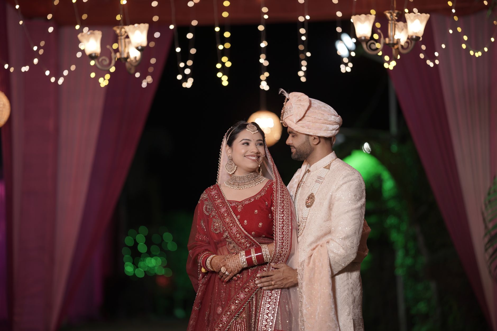 Beautifully dressed Indian bride and groom pose under twinkling lights during a nighttime wedding ceremony